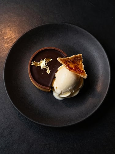 Overhead of a composed dessert plate on matte black ceramic — dark chocolate tart with gold leaf, quenelle of crème fraîche ice cream, caramelised tuile shard — studio strobe balanced with warm ambient light, deep shadow surrounds, cream and gold tones