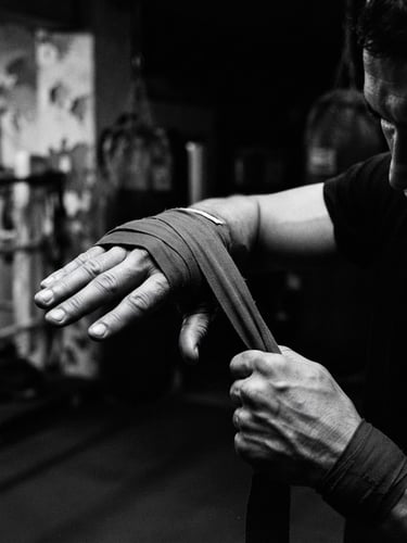 Gritty black and white close-up of a fighter wrapping their hands with red wraps in a dimly lit gym, dramatic side lighting, 35mm film.