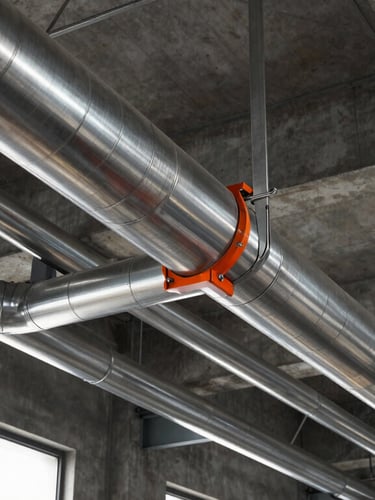 Low-angle sharp industrial photography of clean stainless steel ductwork running across a high concrete factory ceiling, dramatic side lighting cutting through windows, cool steel tones with safety orange accents, 35mm lens.