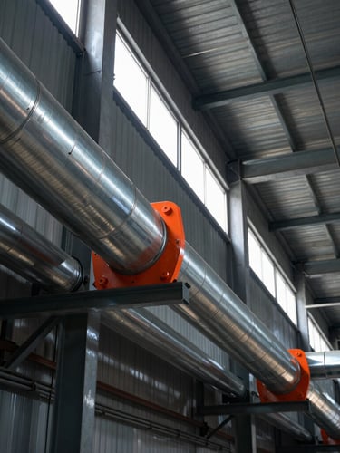 Low-angle linear perspective of heavy-duty stainless steel ductwork running across an industrial factory ceiling, sharp technical details, dramatic side lighting from high windows, cool steel tones with safety orange accents.