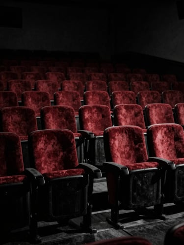 Low-key high-contrast black and white shot of empty vintage cinema seats in a grand old theater, dramatic key light casting long shadows across the red velvet, mysterious atmosphere