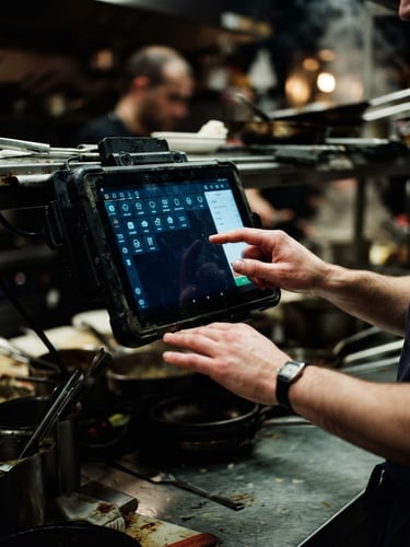 Close-up of a rugged Android POS tablet mounted in a busy, dimly lit kitchen, hands interacting with the screen, high-contrast moody lighting with cool screen glow, rich dark tones.