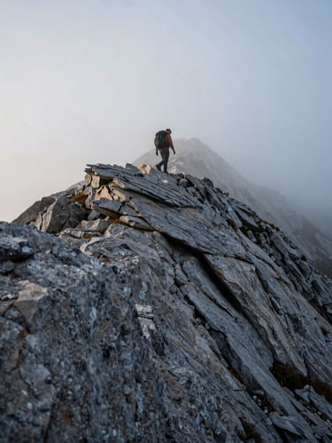 Low-angle wide shot of a lone hiker traversing a rugged, misty mountain ridge at dawn, cold overcast light, sharp granite rocks in foreground