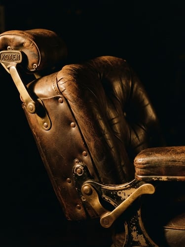 Close-up of a vintage leather barber chair in a low-lit room, dramatic side-lighting, warm amber highlights, deep shadows