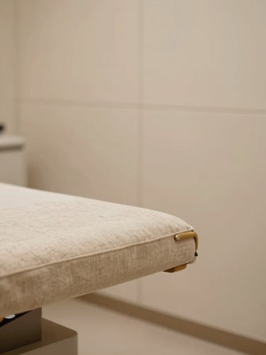 Close-up interior of a refined beauty clinic treatment room — warm diffused light falling on a beige linen headrest, a sleek brushed-gold detail on a treatment chair arm, soft shadow on cream-toned wall panels; no people, no equipment labels, composed to the left third of frame with generous negative space to the right