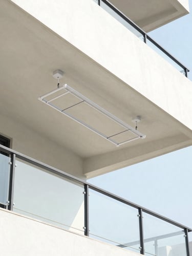 Bright architectural photography of a clean modern apartment balcony in Mysuru, a sleek ceiling-mounted clothes drying system hoisted high against the sunlit concrete ceiling, natural daylight, sharp focus.