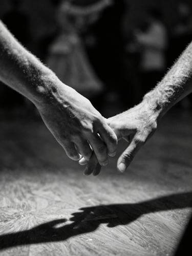 Low angle dramatic shot of hands connecting in ballroom dance, stark spotlight cutting through deep shadows, motion blur on the background, high contrast black and white.