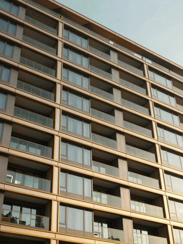 Premium architectural photography of a modern residential building facade, warm golden-hour sunlight reflecting off clean glass panels and champagne gold metal louvers, wide-angle lens