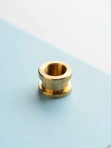 A close-up of a high-tolerance machined brass component resting on a clean, pastel blue surface under soft, diffused daylight, minimalist product photography style