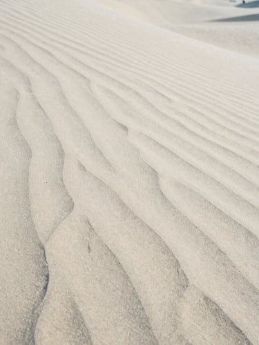 A close-up photograph of wind-swept sand dunes with sharp shadows, soft natural morning light, minimalist composition, medium format film texture.
