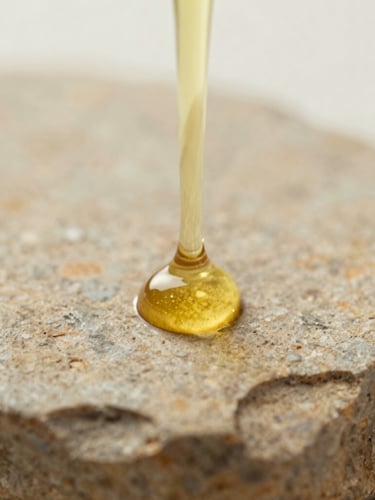Extreme close-up of a single golden drop of cold-pressed sweet almond oil suspended on a textured warm stone surface, soft diffused morning light, shallow depth of field