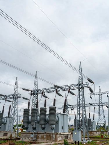 Wide environmental shot from ground level looking up at a large power transformer yard, heavy-gauge cables and steel lattice towers receding into distance, overcast equatorial daylight, no people, organized industrial scale