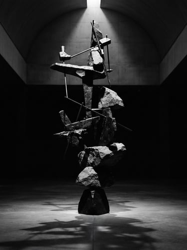 Stark monochrome gallery photography, dramatic chiaroscuro spotlighting, a towering balancing sculpture of bronze and stone in an empty high-ceilinged museum hall, deep obsidian shadows, 35mm