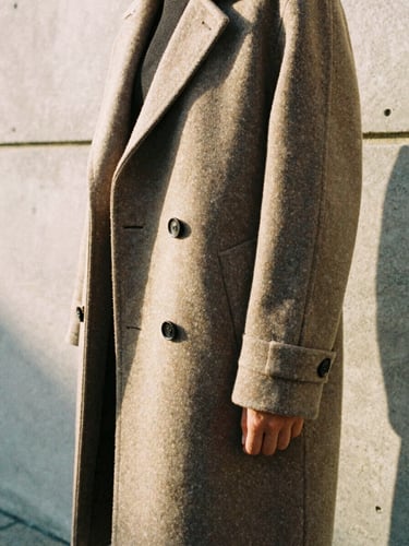 A close-up of a structured wool coat drape against a concrete gallery wall, warm afternoon sun casting soft shadows, 35mm film aesthetic