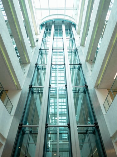 Low-angle wide shot looking up a sleek glass elevator shaft rising through a sunlit corporate atrium, cool teal reflections, bright high-key lighting, architectural symmetry.