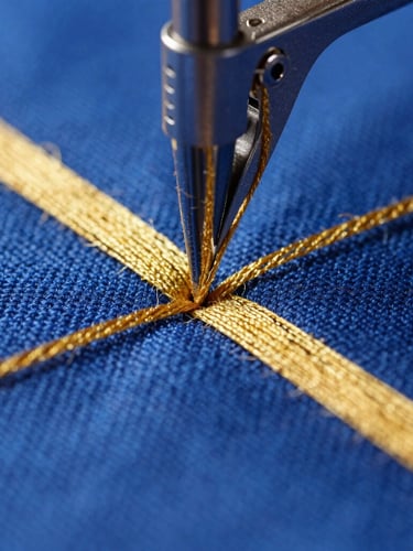 Extreme macro close-up of a high-speed embroidery needle piercing deep royal blue canvas, showing high-density gold thread texture and zero-fray tension under bright studio light.