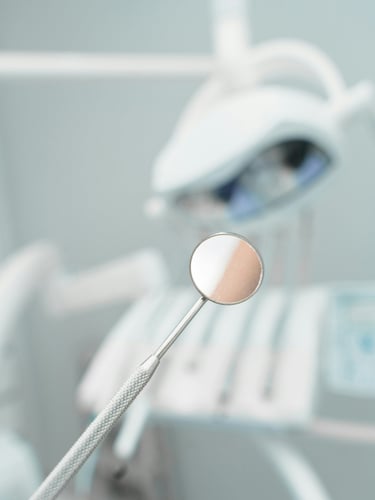 A stainless steel dental mirror held in focus within a modern dental office setting.