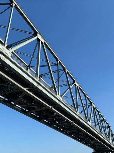 Low-angle sharp architectural photography of a massive steel bridge structure under bright high-contrast daylight, deep blue sky, metallic steel tones