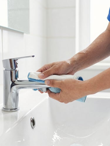 Close-up of a professional cleaner in a neat uniform polishing a chrome bathroom faucet, bright natural daylight, sparkling clean white tiles, sharp focus