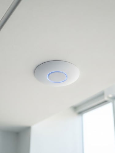 A clean ceiling-mounted UniFi access point glowing with a soft blue LED ring, installed flush on a crisp white ceiling, bright natural daylight, architectural detail.