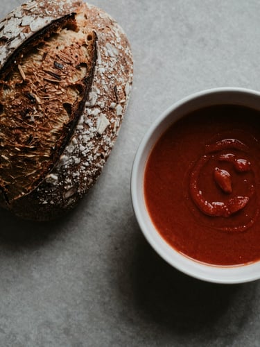 Overhead flat-lay of a rustic sourdough bread next to a ceramic bowl of dark red tomato soup, soft north-facing daylight, moody shadows, editorial food styling