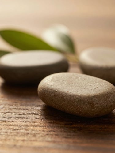 Macro shot of warm massage stones resting on a dark wood surface beside soft green leaves, diffused golden-hour lighting