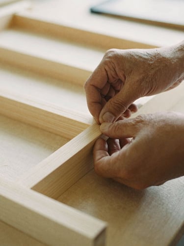 A close-up documentary shot of hands assembling a wooden frame in a bright vocational workshop, natural light, warm organic tones