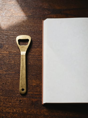 Overhead flat-lay of a brass bottle opener and a heavy paper journal on a dark oak desk, warm morning window light, soft shadows, 35mm photography
