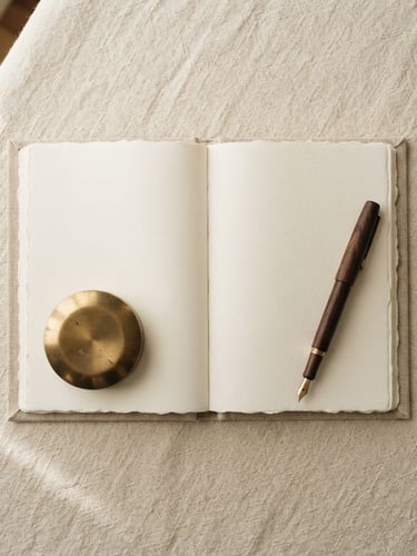 Overhead flat-lay of a brass paperweight, an open linen notebook, and a dark wooden pen resting on a textured cream linen tablecloth, soft morning window light, gentle shadows, warm editorial photography.