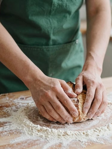 Close-up of hands kneading rustic sourdough on a flour-dusted wooden counter, soft natural side daylight, matte forest green apron visible, 35mm film.