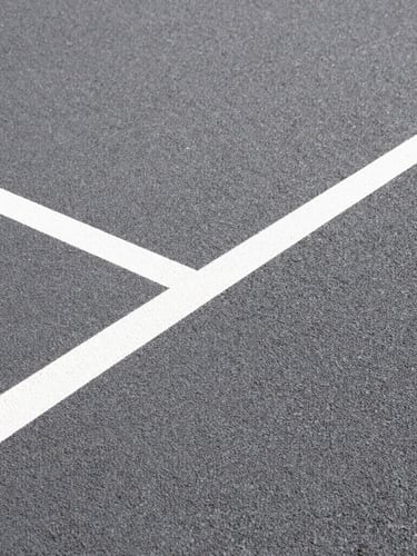 Extreme close-up of textured acrylic court surface grain, sharp geometric white line cutting diagonally, direct bright sunlight showing high-contrast texture.