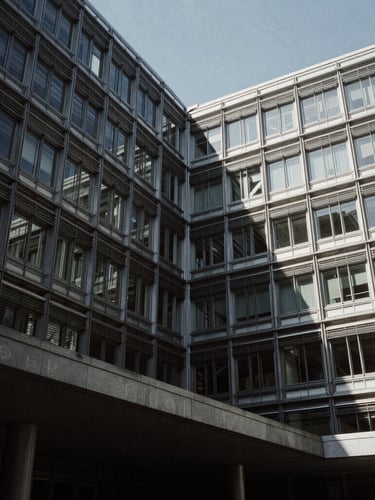 Low-angle architectural shot of a brutalist steel and glass government ministry under harsh directional light, deep shadows, desaturated cool tones.