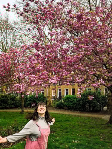 Laura dances in front of a pink blossom tree in London. she is wearing pink overalls and smiling.