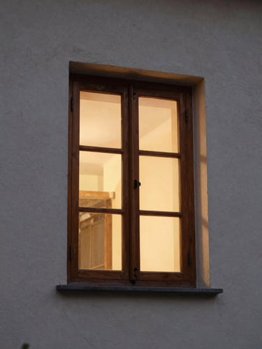 Tight architectural crop of a tall open wooden window at dusk, warm interior light spilling onto a textured plaster wall, soft shadows, editorial photography.