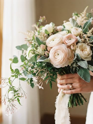 A close-up shot of an asymmetrical bridal bouquet with delicate blush ranunculus, trailing jasmine vines, and textured sage foliage, held gently in soft morning window light, 35mm photography