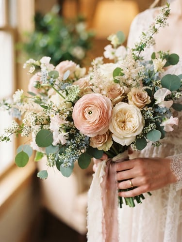Close-up of a delicate asymmetrical bridal bouquet with blush ranunculus and pale green eucalyptus held naturally in soft morning window light, shallow depth of field, 35mm