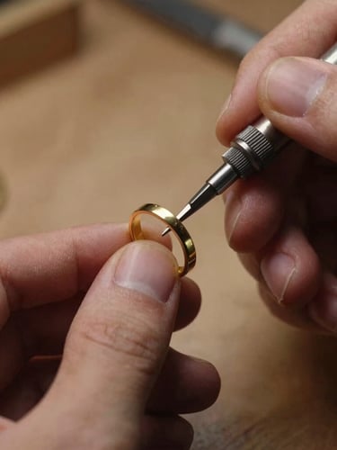Macro close-up of a jeweler's hands using a traditional engraving tool on a solid gold ring, warm side-lighting, soft shadows, workshop background.