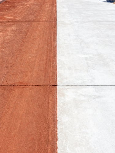 Before-and-after split view of a weathered concrete driveway in Broome, left side covered in thick red Kimberley dirt, right side blasted clean to pristine white concrete under harsh afternoon sun, 35mm