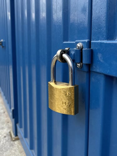High-contrast industrial lighting on a row of clean, blue corrugated steel storage doors, a heavy-duty brass padlock secured on the latch in the foreground, sharp geometric perspective down the concrete aisle.