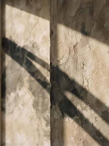 Wide, minimalist editorial photograph of a textured plaster wall with soft, long shadows from an unseen window, muted warm sand and charcoal tones, shallow depth of field, abstract architectural detail.
