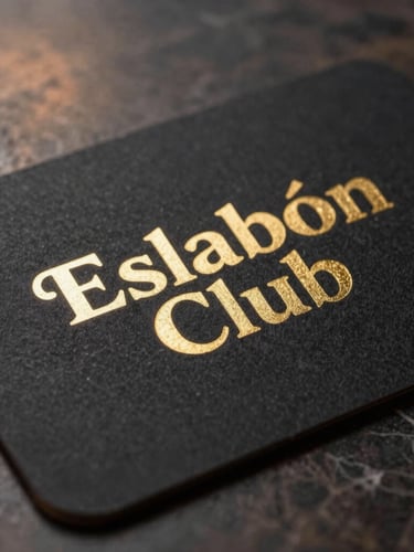 Macro close-up of a matte black, heavy paper card with the gold-embossed Eslabón Club emblem, resting on a dark marble surface, warm side lighting