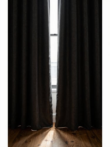 Low-key moody editorial photo of heavy charcoal blackout curtains framing a quiet window, deep shadows, warm oatmeal light reflecting off a wooden floor, 50mm lens