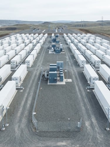 Wide high-angle technical photograph of a utility-scale battery energy storage system installation, clean white containerized battery units, gravel ground, bright overcast daylight, cool slate and blue tones, sharp architectural focus.