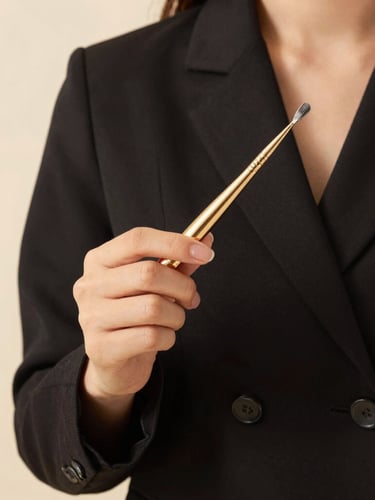 An editorial close-up of a student in a chic black uniform holding a premium gold-accented applicator tool under warm, diffused studio lighting, 35mm