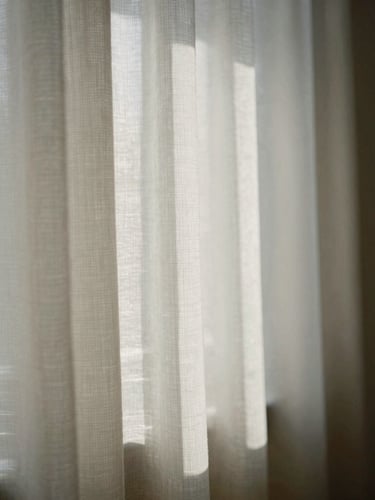 A portrait crop of soft diffused morning light filtering through structured linen fabric, casting gentle shadows, quiet studio corner, minimalist composition