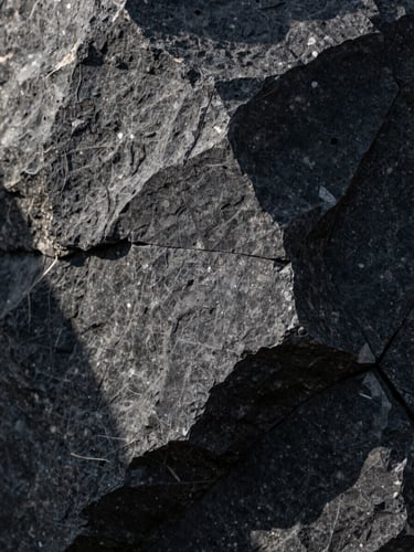Close-up macro shot of dark basalt stone with sharp, natural geometric cleavage, directional morning light casting deep shadows, architectural texture.