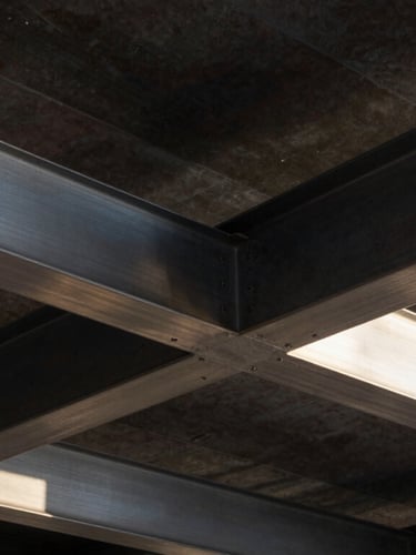 Close-up architectural photography of premium construction materials, brushed steel beams meeting dark structured concrete, sharp afternoon sunlight casting geometric shadows.