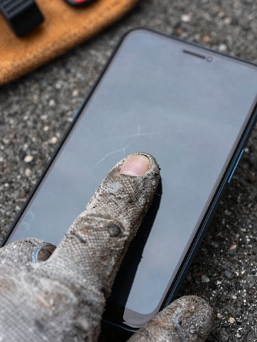 Extreme close-up from above, a gloved thumb pressing a cracked-screen Android phone, overcast outdoor light, worn work glove texture sharp in foreground, device screen faintly visible beneath thumb, asphalt and a tool belt edge out of focus in background
