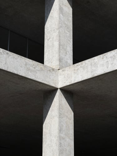 Portrait-oriented architectural photograph of a detailed structural join between concrete and steel, captured in harsh, directional sunlight, deep shadows highlighting texture.