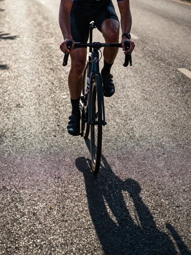 Low-angle hero shot of a cyclist in intense effort, sweat on skin, early morning light casting long dramatic shadows on the asphalt